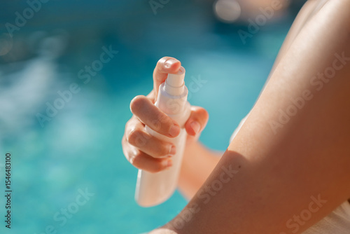 Close-up of a woman spraying sunscreen on her arm by the pool, highlighting sun protection and summer skincare.