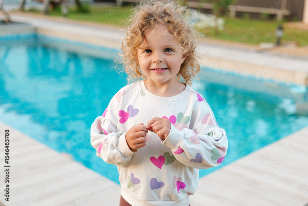 Naklejka premium Happy curly-haired girl in heart-patterned sweatshirt smiling by the pool on a sunny day, enjoying summer fun and outdoor leisure.