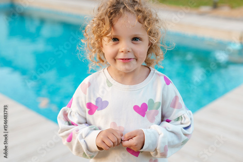 Smiling curly-haired girl in heart-print sweatshirt stands by the pool, enjoying a warm summer day with a playful and innocent expression.