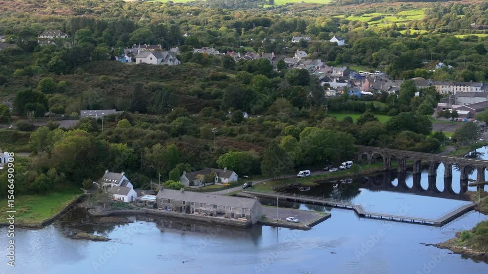 An overhead view showcases the calm river reflecting a stone bridge and nearby waterfront structures. Homes are scattered amidst lush green trees and hills. An overhead view showcases the calm river reflecting a stone bridge and nearby waterfront structures. Homes are scattered amidst lush green trees and hills.