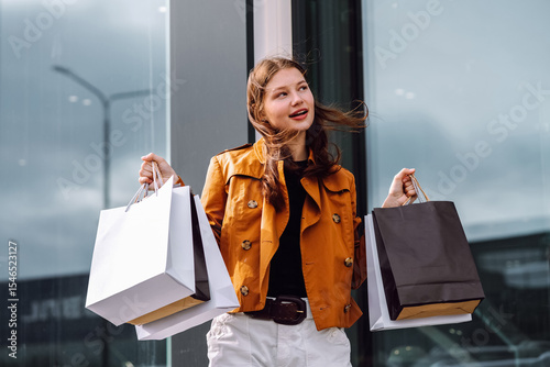 Young woman celebrates after shopping outdoors with bags in hand on a sunny day in an urban setting. Consumerism, purchases, shopping, lifestyle, sale concept.