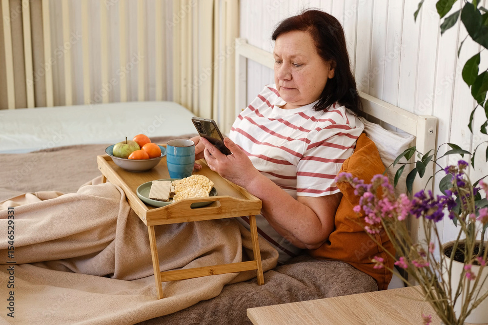 Senior woman having breakfast sitting in bed, drinking tea, enjoying her morning at home. Old lady eating from tray and using phone. Elderly female in pajamas relaxing with cup of coffee in a bedroom. Senior woman having breakfast sitting in bed, drinking tea, enjoying her morning at home. Old lady eating from tray and using phone. Elderly female in pajamas relaxing with cup of coffee in a bedroom.
