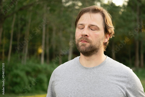 Portrait of handsome young man in gray t-shirt outdoors. Male with long hair and beard feel happy, calm and serious on Green nature sunset background. Man looking away into distance. Diversity People