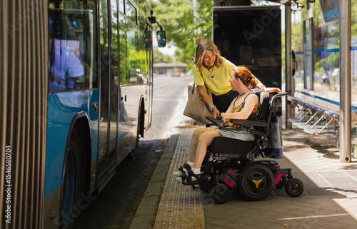 Woman helping disabled girl on wheelchair to board bus at bus stop
