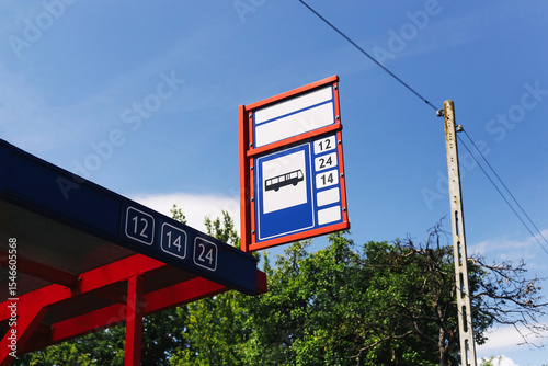 Bus stop sign with route numbers. Public transport station under blue sky. Urban travel infrastructure in city suburb. Red and white signage with trees in background.