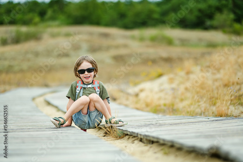 A cheerful child wearing sunglasses and a backpack running playfully on a wooden boardwalk at the Curonian Spit in Lithuania, enjoying an outdoor adventure.