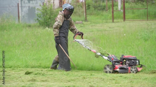 A man mows the grass with a lawnmower. A man in a safety suit and mask is mowing long grass with a gas-powered lawn mower.