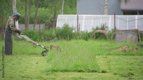 A man mows the grass with a lawnmower. A man in a safety suit and mask is mowing long grass with a gas-powered lawn mower.
