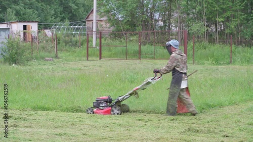 A man mows the grass with a lawnmower. A man in a safety suit and mask is mowing long grass with a gas-powered lawn mower.