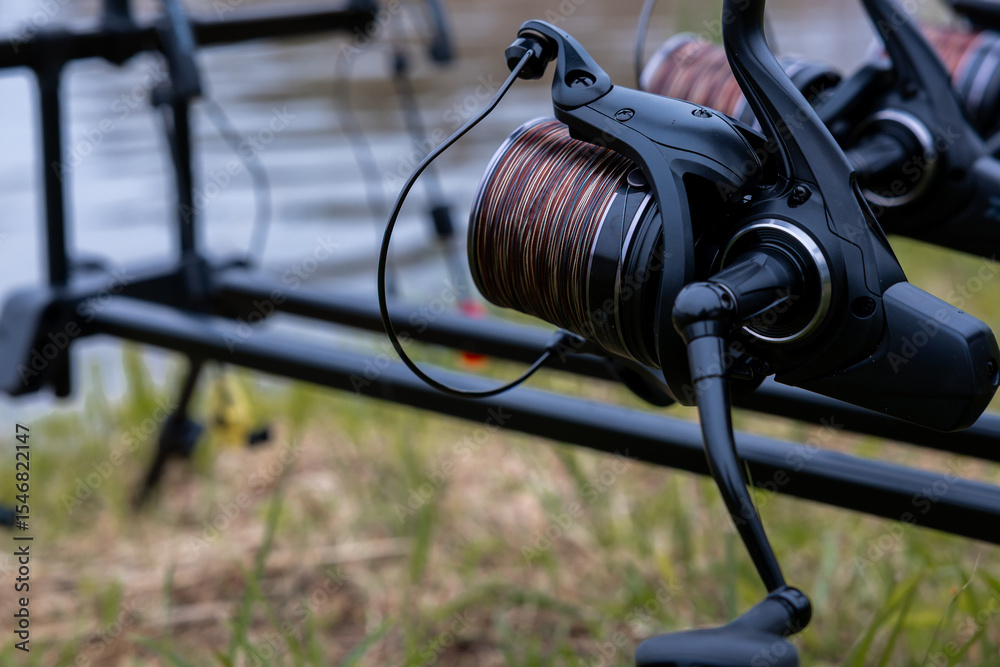 Fishing reel mounted on a rod holder with tightly wound line, positioned near water. Grass is visible in the foreground, adding natural detail. Fishing reel mounted on a rod holder with tightly wound line, positioned near water. Grass is visible in the foreground, adding natural detail.