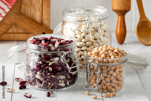 Variety of uncooked beans and chickpeas stored in glass jars on bright wooden surface in rustic kitchen setting