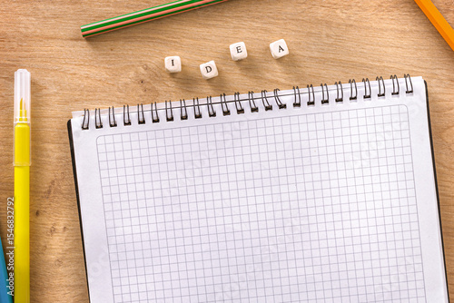 Top view of empty squared notebook with colorful pencils and wooden cubes forming the word idea on desk background
