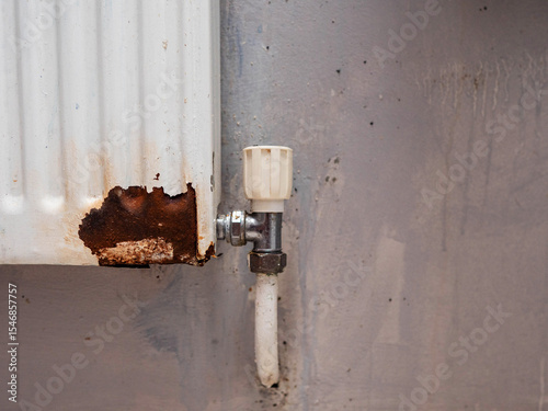 A rusty radiator with a white cap. The radiator is old and has been exposed to the elements for a long time, the wall behind is grey and grungy, Budget or cheap flat. Low income or lack of funds
