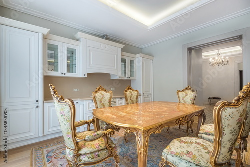 The refined interior of a luxurious dining room kitchen in a classic style. White kitchen set, inlaid table and chairs with curved legs, gilt and silk upholstery.