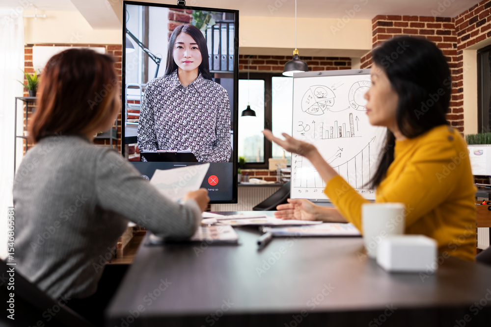 Fototapeta premium Company advisors seated at desk with documents, shares business updates with a remote client via video call on led screen. Asian women discussing project analysis during virtual meeting with manager.