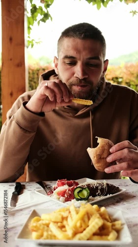 Man holding bread and picking up fries while eating a traditional Serbian meal with grilled meat and vegetables. Vertical 4k footage