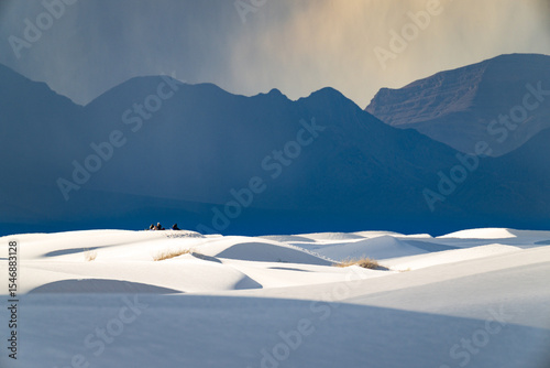 White rolling sand dunes and dark mountains in the distance under a rain cloud at White Sands National Park, New Mexico. A group of hikers can be seen in the distance.
