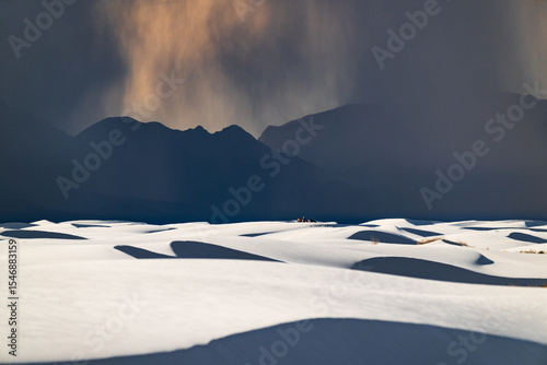 White rolling sand dunes and dark mountains in the distance under a rain cloud at White Sands National Park, New Mexico. A group of hikers can be seen in the distance.