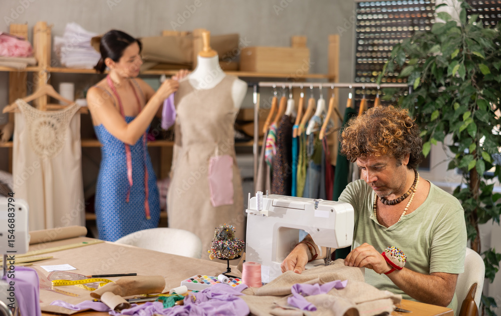 Fototapeta premium Focused middle-aged tailor using sewing-machine and tailoress working with dummy in workshop