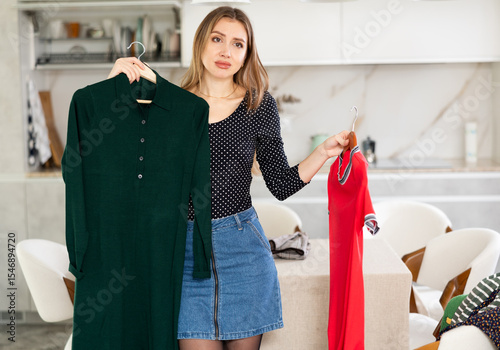 Upset young woman cannot choose a dress for the holiday. Difficult choice
