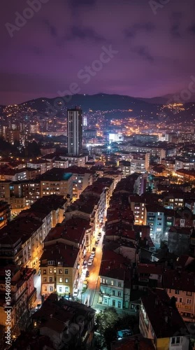 High-angle night view of a vibrant city with illuminated buildings and mountains in the distance, timelapse
