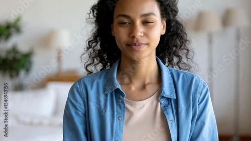 A Young smiling woman with curly hair looks directly at the camera in a bright living space while wearing a denim shirt.