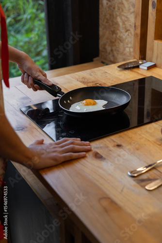 Photo of a young woman gripping the frying pan handle while frying an egg, showing a relaxed cooking moment in a cozy kitchen with natural light highlighting the fresh food

