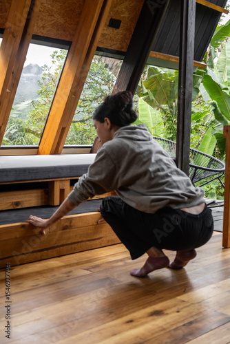 A young woman is seen from behind as she squats inside a rustic wooden cabin. Her posture suggests focus and calm, blending with the warm and peaceful atmosphere of this quiet mountain retreat.

