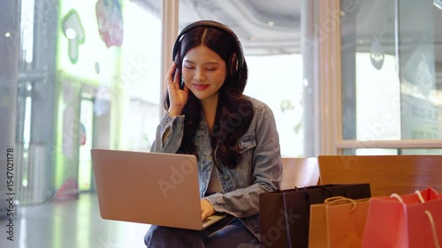 Inside a urban cafe, happy young asian woman wearing wireless headphones smiles as she selects her go-to energetic disco tracks on a laptop. The relaxed atmosphere inspires her to dance joyfully