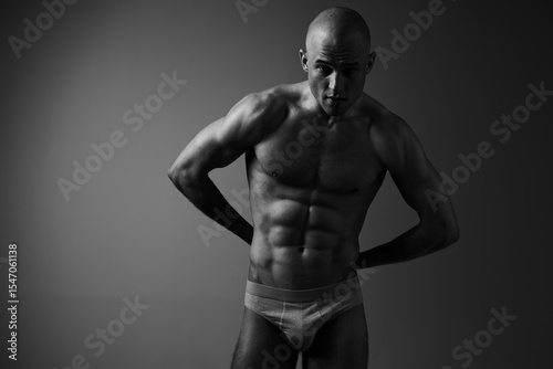 Black and white image of a muscular man posing confidently, showcasing his toned physique and athleticism in a dramatic light setting.