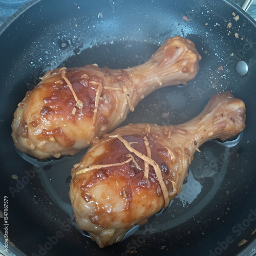 Two glazed chicken drumsticks simmer gently in a black frying pan ready to be served for dinner tonight.
