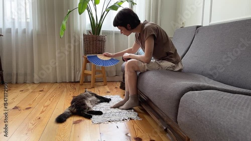 Tired caring woman fan overheating cat on floor in hot apartment with no AC. Pet owner struggle in summer in non ventilated room, struggle with high temperatures, indoor discomfort, pet care priority