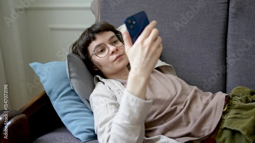 Relaxed woman browsing social media on smartphone while lounging on sofa, absorbed in digital web feed. Device dependency, endless scrolling habit, internet downtime, modern leisure, lonely isolation