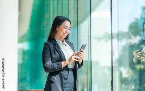 Asian businesswoman using smartphone near glass wall with reflection. Confident woman in formal outfit using phone in modern indoor space.
