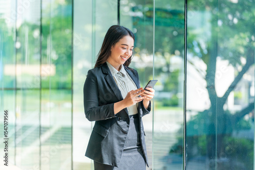 Asian businesswoman using smartphone near glass wall with reflection. Confident woman in formal outfit using phone in modern indoor space.