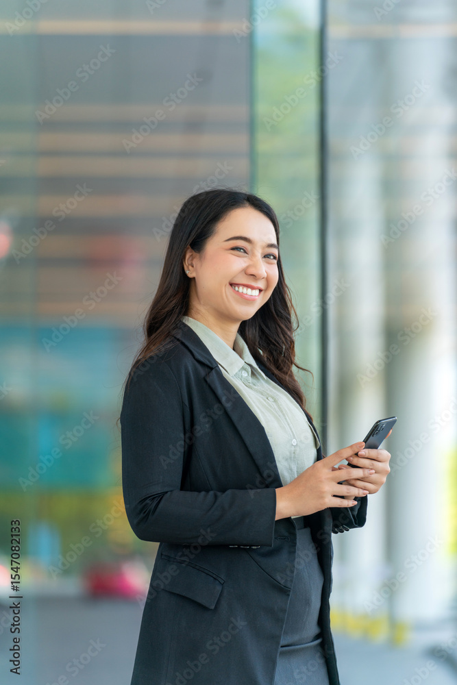 Smiling Asian businesswoman using smartphone indoors near glass wall. Confident professional woman holding phone in modern office building. Smiling Asian businesswoman using smartphone indoors near glass wall. Confident professional woman holding phone in modern office building.
