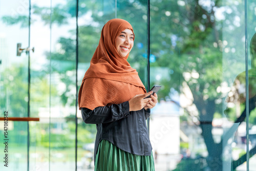 Asian muslimah using mobile phone with cheerful expression in bright modern space. Happy Muslim woman holding smartphone near glass window with greenery outside.