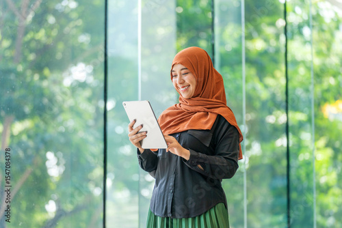 Smiling Asian muslimah using tablet indoors with natural outdoor view. Confident Muslim woman working on digital device near glass window.
