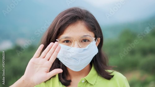4K Portrait of young Indian woman wearing mask and weaving hand to say hello, hi. Female tourist wearing mask to protect herself from flue, corona virus and pollution. Health care and safety concept