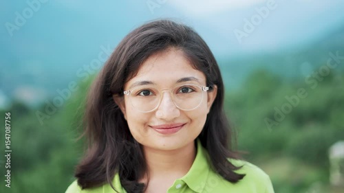 Smiling modern Indian girl starring at camera with happy smiling face while standing against mountains in Manali, India. Happy cheerful teenager girl portrait. Female with positive emotions.