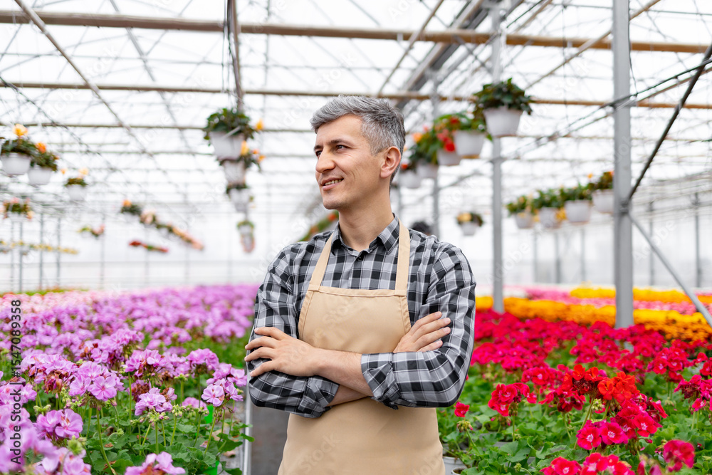 Obraz premium A smiling man in a greenhouse, surrounded by colorful flowers, wearing an apron and plaid shirt, looking content.