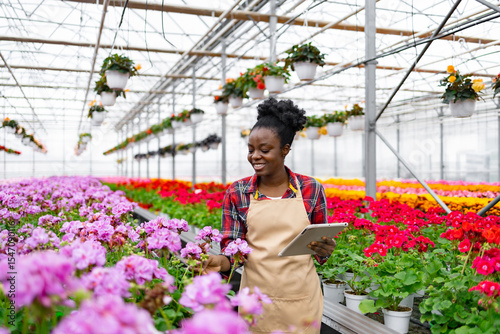 An African-American woman tends to flowers in a greenhouse, using a tablet to monitor growth and development.