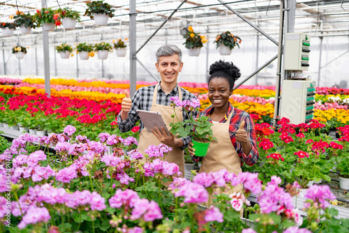 Two smiling people in a greenhouse with flowers, giving thumbs up. One holds a tablet, the other a plant.