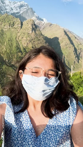 4K Vertical selfie shot of Indian teenager girl wearing mask and standing in mountains. Healthcare, wellness and precaution concept. Corona virus pandemic safety. Girl travel, wear mask