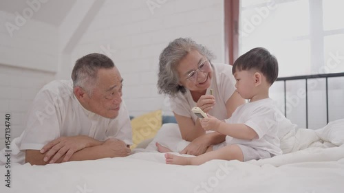 Grandparents joyfully engage with their grandson while he explores a smartphone in a cozy bedroom setting