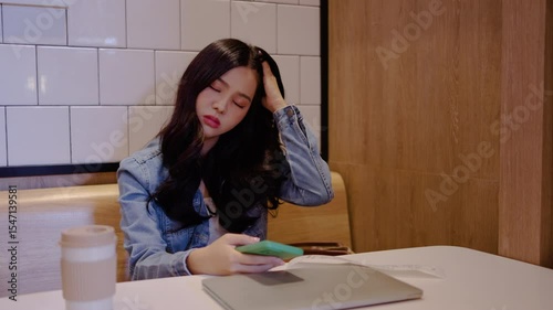 In a restaurant, young asian woman sits alone, visibly stressed as she reveals her bills and calculates expenses on her phone, striving to manage her finances effectively.