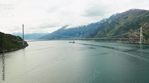 Hardanger Bridge gracefully arches over fjord calm waters with sailing sizable cruise liner. Majestic mountains encapsulates serene beauty of Norway