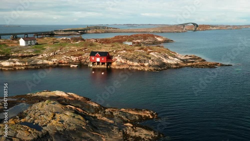 Charming red boathouse on rocky islet reflecting in calm waters by Norwegian coast. Iconic Atlantic ocean road gracefully connects distant islands