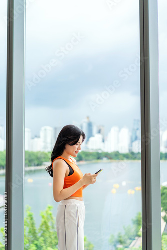 Young Asian woman using smartphone near large window with city view. Casual businesswoman using phone indoors. Free Download Young Asian woman using smartphone near large window with city view. Casual businesswoman using phone indoors. - GFXLoad