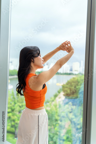 Free Download Young Asian woman standing by large window with city view. Confident modern woman in casual sportswear posing indoors. - GFXLoad